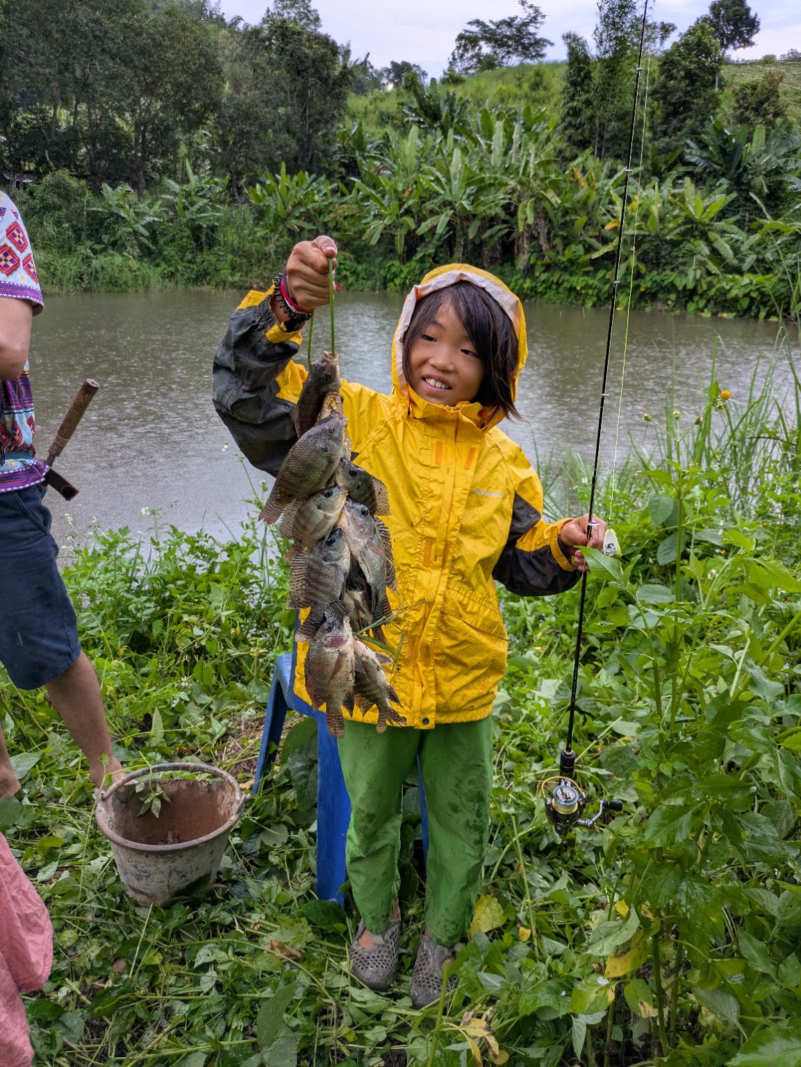 池のそばで、釣った魚を手に笑う黄色い雨具の長男